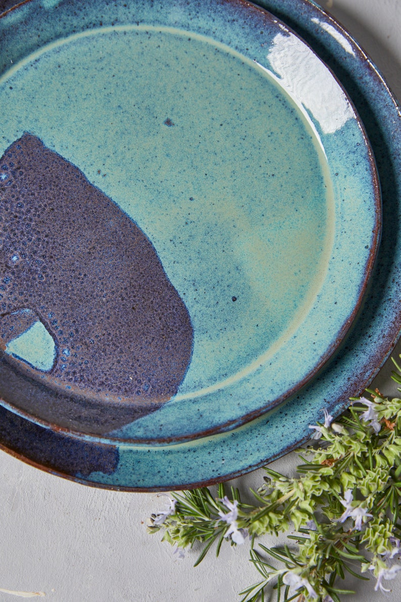 May include: Two ceramic plates, one stacked on top of the other. The top plate is a light blue with a dark blue and brown splatter pattern. The bottom plate is a darker blue with a brown splatter pattern. The plates are on a white surface with green rosemary sprigs.