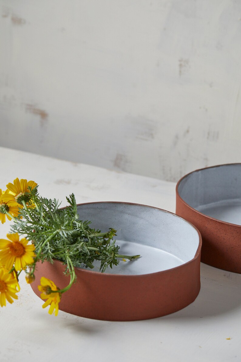 May include: Two terracotta ceramic bowls with a white glaze inside. The bowl on the left has a bouquet of yellow flowers inside.