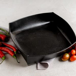 May include: A black square ceramic serving dish with rounded corners. Red chili peppers, cherry tomatoes, and rosemary sprigs are arranged around the dish on a gray cloth, creating a vibrant food presentation.