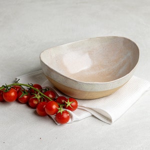 May include: A white ceramic bowl with a speckled glaze sits on a white linen napkin. A sprig of red cherry tomatoes is next to the bowl.