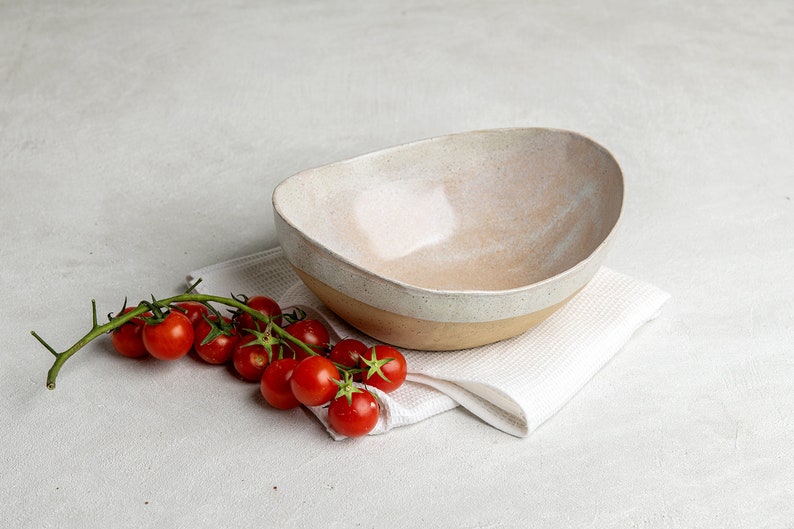 May include: A white ceramic bowl with a speckled glaze sits on a white linen napkin. A sprig of red cherry tomatoes is next to the bowl.