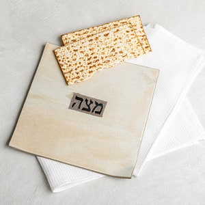 May include: A still-life image featuring matzah crackers atop a beige serving board with Hebrew text, resting on a white textured cloth. The scene is set against a light gray background, evoking a sense of simplicity and tradition.