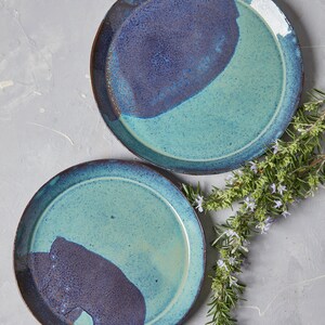 May include: Two ceramic plates with a blue and purple glaze. The plates are round and have a slightly raised rim. The plates are on a light grey background.