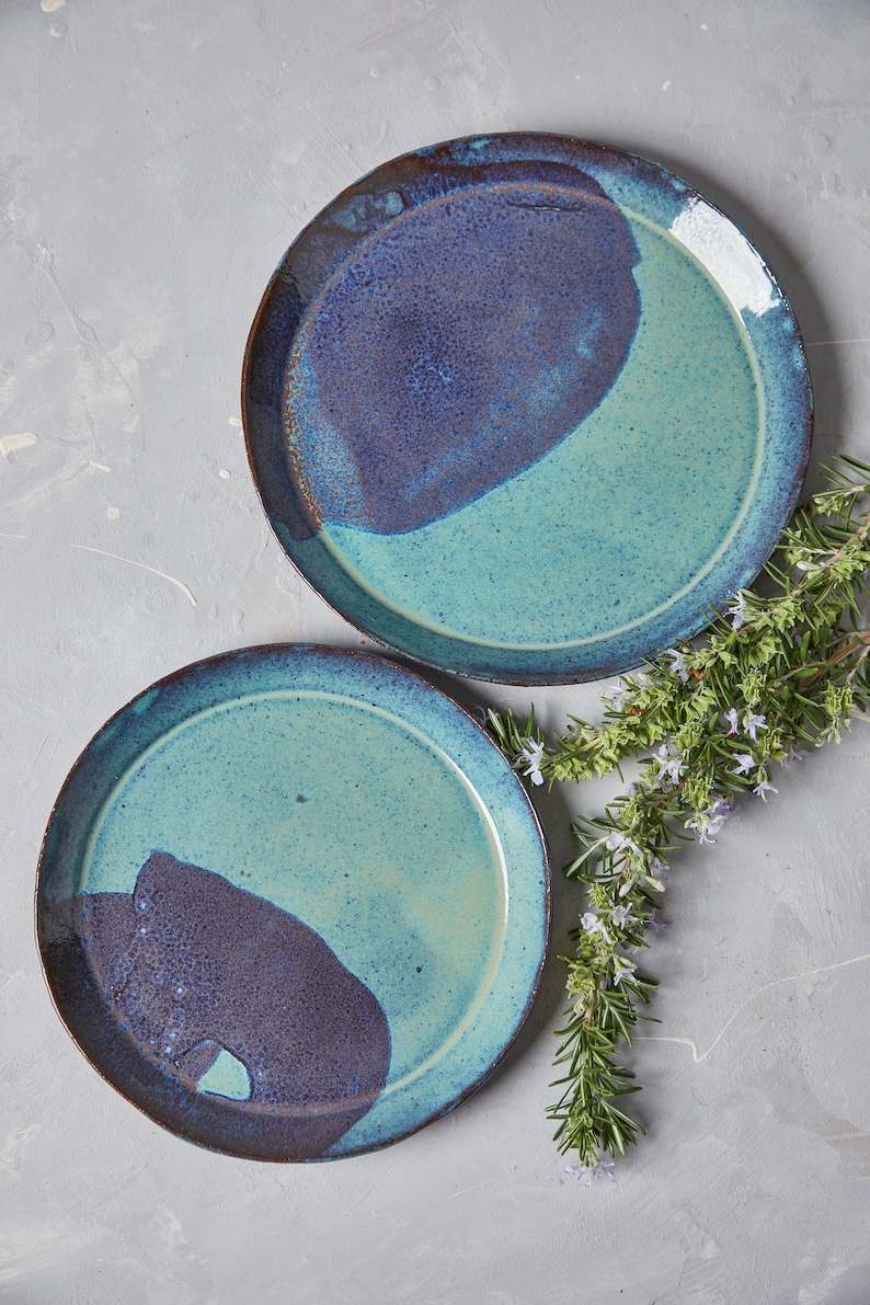May include: Two ceramic plates with a blue and purple glaze. The plates are round and have a slightly raised rim. The plates are on a light grey background.