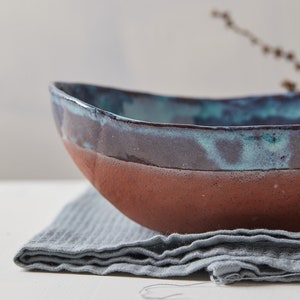May include: A large, oval-shaped ceramic bowl with a teal and brown glaze. The bowl is sitting on a gray linen napkin.