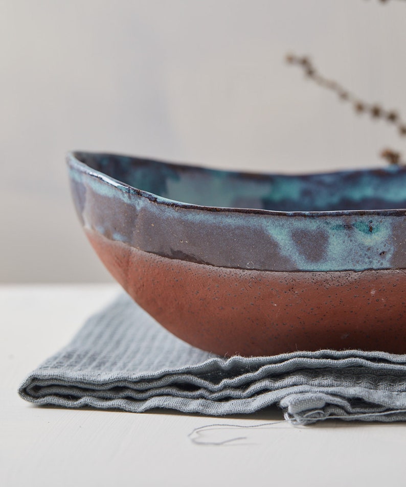May include: A large, oval-shaped ceramic bowl with a teal and brown glaze. The bowl is sitting on a gray linen napkin.