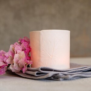 Pode incluir: Um porta-velas de cerâmica branca com um design de folha. O porta-velas está sobre uma toalha de linho cinza com flores de buganvília rosa.