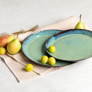 May include: Two turquoise ceramic plates with a dark blue rim are displayed on a beige cloth, alongside golden forks, pears, and small yellow lemons. The plates have a rustic, handcrafted appearance, perfect for serving food.