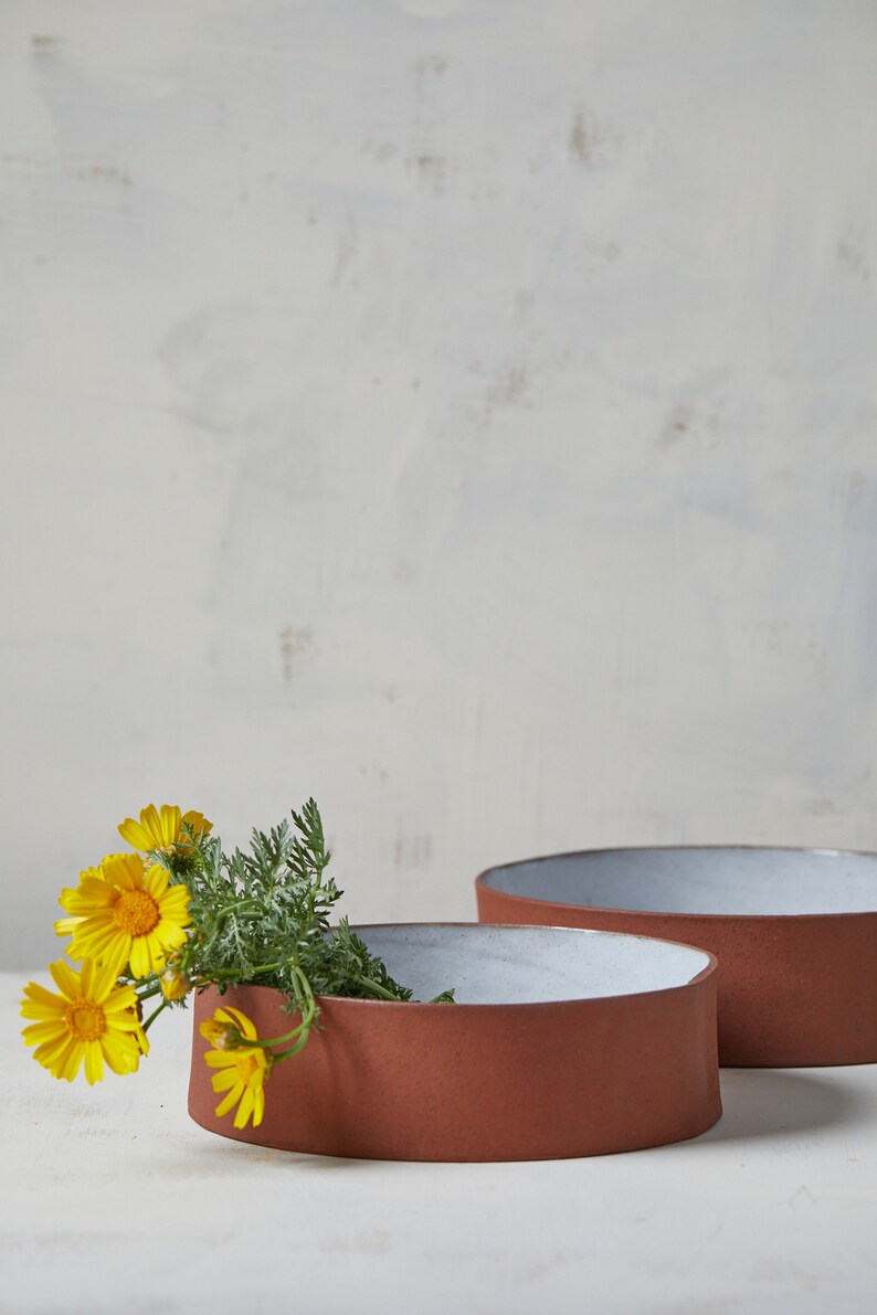 May include: Two terracotta bowls with white glaze interiors. The smaller bowl is filled with a bouquet of yellow wildflowers.