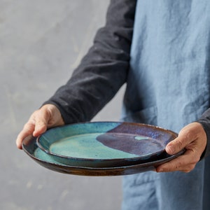 May include: Two ceramic plates, one stacked on top of the other. The top plate is a light blue-green colour with a dark brown glaze. The bottom plate is a dark brown colour with a light blue-green glaze.