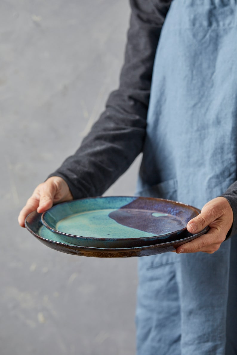 May include: Two ceramic plates, one stacked on top of the other. The top plate is a light blue-green colour with a dark brown glaze. The bottom plate is a dark brown colour with a light blue-green glaze.