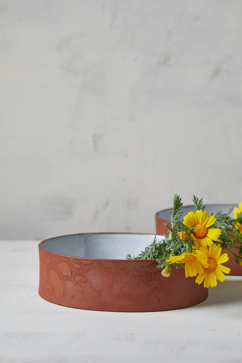 May include: Two terracotta colored ceramic bowls with a white glaze on the inside. A bouquet of yellow flowers with green leaves is placed in the bowl on the right.