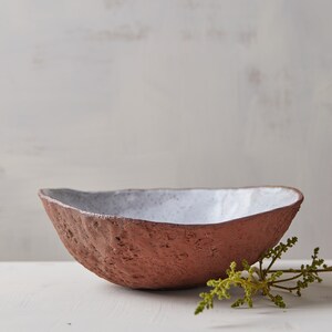 Op de afbeelding: Een witte en terracotta keramische kom met een ruw, getextureerd exterieur. De kom staat op een wit oppervlak met een takje groen blad.