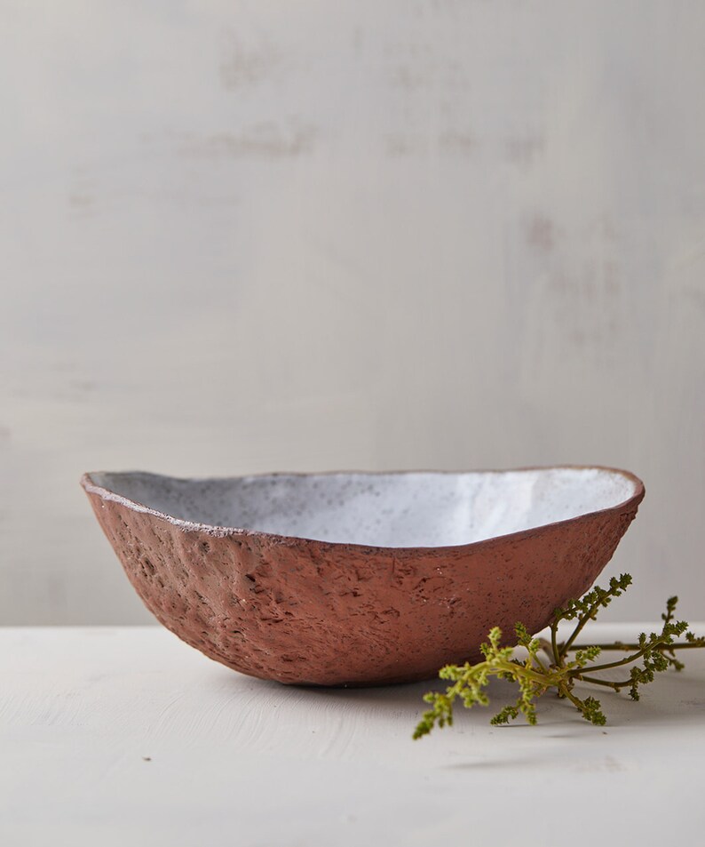 Op de afbeelding: Een witte en terracotta keramische kom met een ruw, getextureerd exterieur. De kom staat op een wit oppervlak met een takje groen blad.