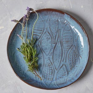 May include: A blue ceramic plate with a fish design. The plate has a brown rim and is decorated with a raised fish pattern. A sprig of lavender is placed on the plate.