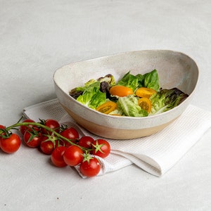 May include: A white ceramic bowl with a light brown rim filled with a green salad with halved yellow cherry tomatoes. A sprig of red cherry tomatoes is on a white linen napkin next to the bowl.