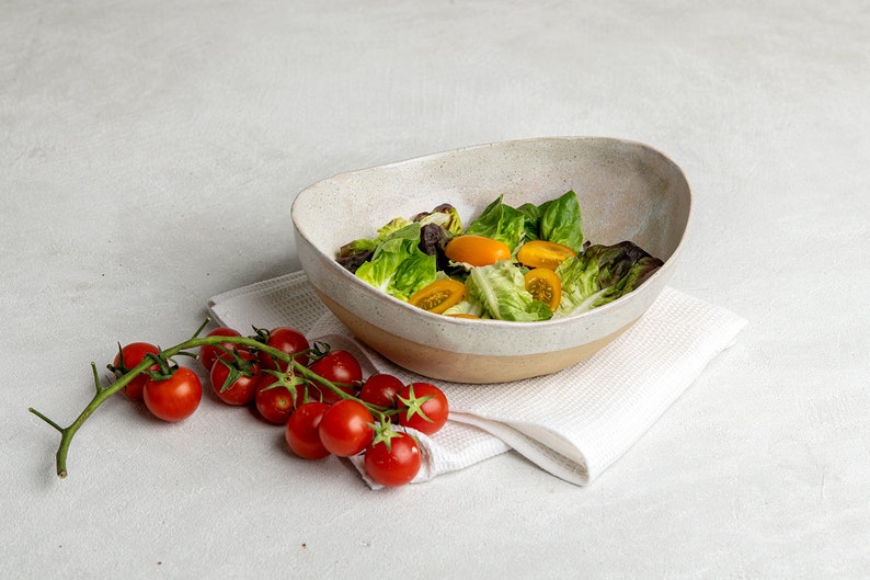 May include: A white ceramic bowl with a light brown rim filled with a green salad with halved yellow cherry tomatoes. A sprig of red cherry tomatoes is on a white linen napkin next to the bowl.