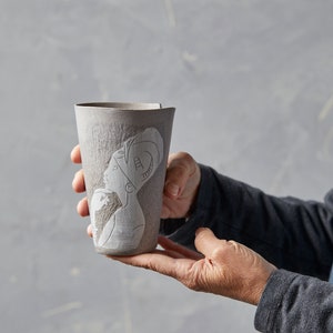 May include: A grey ceramic jug with a white relief design of a woman's face. The jug is being held by a person's hand.