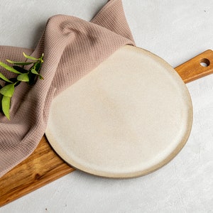 以下が含まれることがあります： 木製のまな板の上に、茶色の縁が付いた丸いオフホワイトのセラミックプレートが置かれています。 モーヴのワッフル織りのタオルと、オリーブの付いた緑の葉の小枝がボードに掛けられており、素朴で自然な美しさを作り出しています。