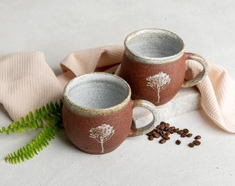 以下が含まれることがあります： 白い木のデザインが施された茶色のセラミック製のマグカップが2つ。マグカップは白い表面に置かれ、その周りにコーヒー豆が散らばっています。