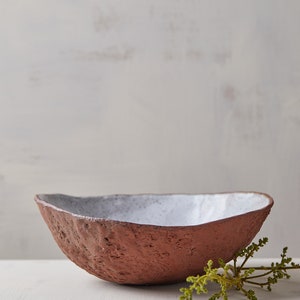 Op de afbeelding: Een witte en terracotta keramische kom met een ruw, getextureerd exterieur. De kom staat op een wit oppervlak met een groene tak van gebladerte.
