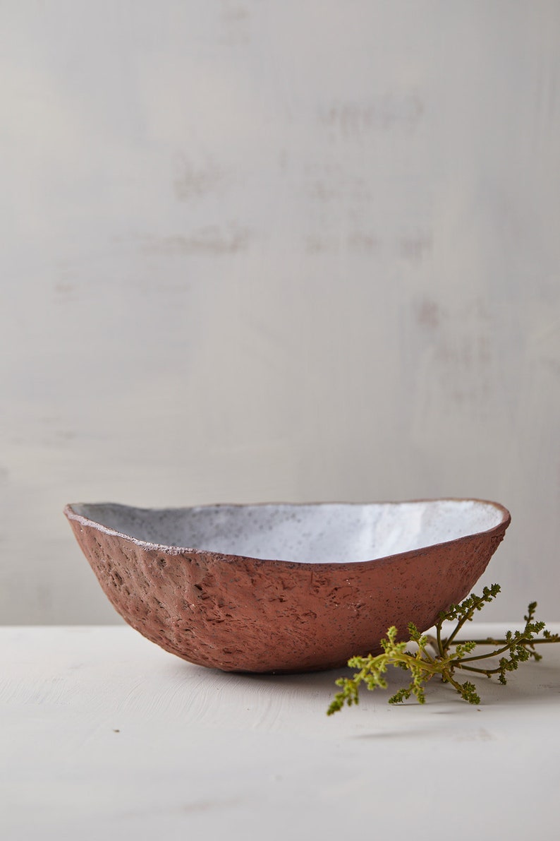 Op de afbeelding: Een witte en terracotta keramische kom met een ruw, getextureerd exterieur. De kom staat op een wit oppervlak met een groene tak van gebladerte.