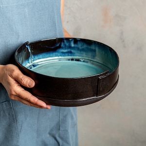 May include: A large, round ceramic bowl with a dark brown exterior and a light blue interior. The bowl has a slightly raised rim and a unique, uneven glaze.