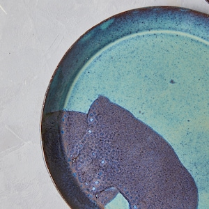 May include: A round ceramic plate with a blue and brown glaze. The plate has a unique design with a dark brown section on one side and a light blue section on the other side.