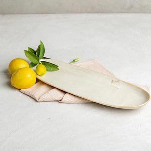 May include: An elongated, oval-shaped ceramic serving platter with a smooth, off-white glazed finish. The platter is positioned next to two bright yellow lemons with green leaves attached, resting on a folded, light pink textured fabric cloth.
