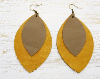 May include: A pair of dangle earrings with two layers of leather. The top layer is a light brown color and the bottom layer is a mustard yellow color. The earrings are shaped like a teardrop.