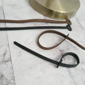 May include: Three brown leather cord ties and one black leather cord tie with a metal buckle. The ties are on a white tiled surface.