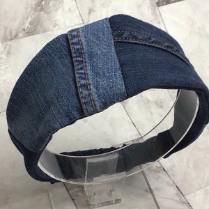 Może przedstawiać: Opaska do włosów z denimu wykonana z odzyskanych niebieskich dżinsów. Opaska jest wykonana z kilku kawałków denimu zszytych ze sobą. Opaska znajduje się na przezroczystym stojaku.