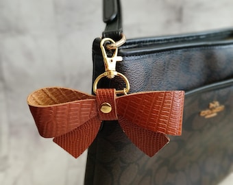 May include: A brown leather bow keychain with a gold metal clasp attached to a black handbag.