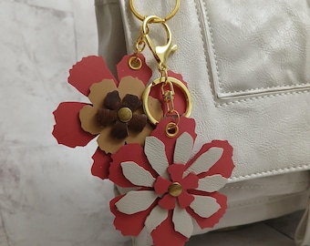 May include: A gold key chain with a red and white flower charm. The charm is made of leather and has a brown center.