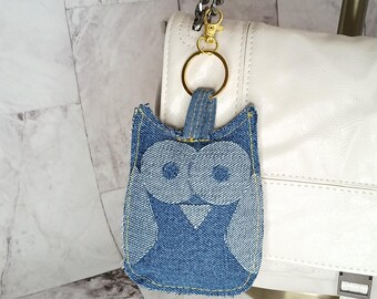May include: A denim owl keychain with large, round eyes and yellow stitching. The owl is attached to a gold-colored keyring and a silver chain. The keychain hangs from a white purse.