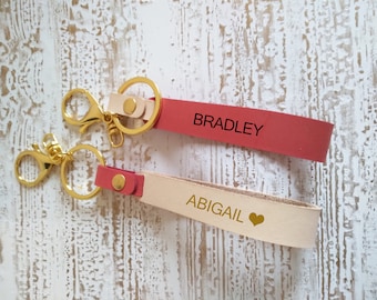May include: Two personalized leather keychains with gold metal key rings. One keychain is pink with the name "BRADLEY" and the other is beige with the name "ABIGAIL" and a gold heart.