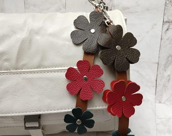 May include: White leather handbag with a chain strap and a decorative flower charm. The charm is made of brown, red, and green leather and features five flowers.