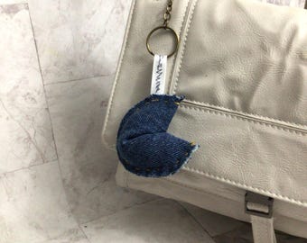 May include: A white leather bag with a denim fortune cookie charm attached to a gold chain. The charm has the word "karma" written on it.