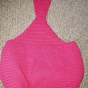 May include: A handmade, bright pink crochet bag with a long handle. The bag has a rounded, open shape, and the handle is a continuation of the crochet work. The texture of the crochet is visible throughout the bag.