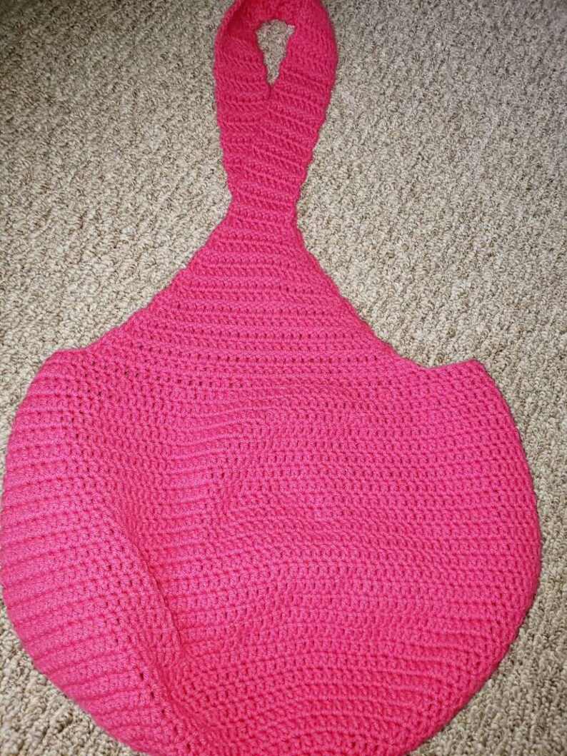 May include: A handmade, bright pink crochet bag with a long handle. The bag has a rounded, open shape, and the handle is a continuation of the crochet work. The texture of the crochet is visible throughout the bag.