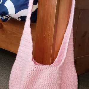 Op de afbeelding: Een lichtroze, gehaakte schoudertas met een lange band. De tas is gemaakt van zacht, getextureerd garen en heeft een afgeronde bodem. De tas hangt aan een houten paal.