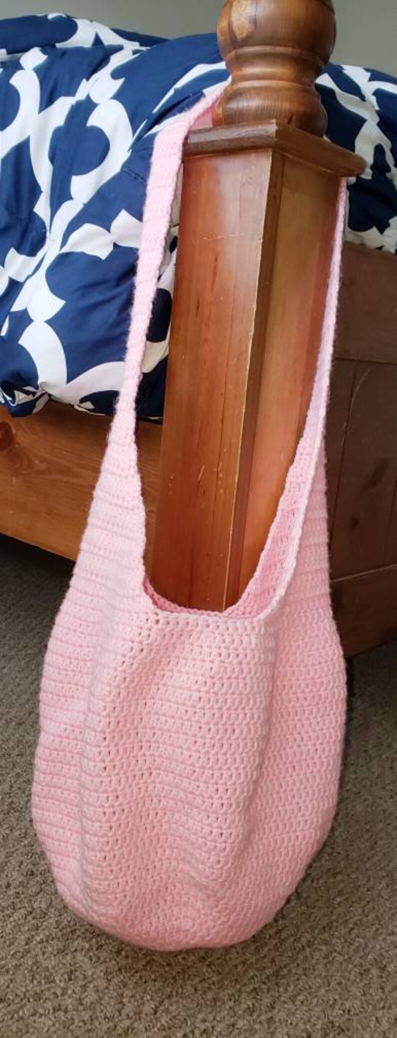 Op de afbeelding: Een lichtroze, gehaakte schoudertas met een lange band. De tas is gemaakt van zacht, getextureerd garen en heeft een afgeronde bodem. De tas hangt aan een houten paal.