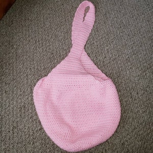 Op de afbeelding: Een lichtroze, handgemaakte gehaakte tas met een lange, geluste handgreep. De tas heeft een afgeronde bodem en een getextureerd oppervlak dat is gemaakt door de haaksteken. De tas is gemaakt van zacht garen.