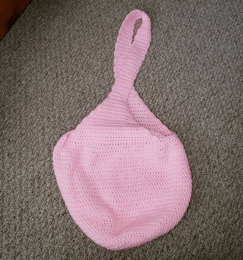 Op de afbeelding: Een lichtroze, handgemaakte gehaakte tas met een lange, geluste handgreep. De tas heeft een afgeronde bodem en een getextureerd oppervlak dat is gemaakt door de haaksteken. De tas is gemaakt van zacht garen.