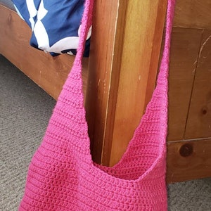 May include: A bright pink, handmade crochet shoulder bag. The bag is a solid color and has a textured appearance. The strap is also crocheted. The bag is shown resting on a carpeted floor next to a wooden bed frame.