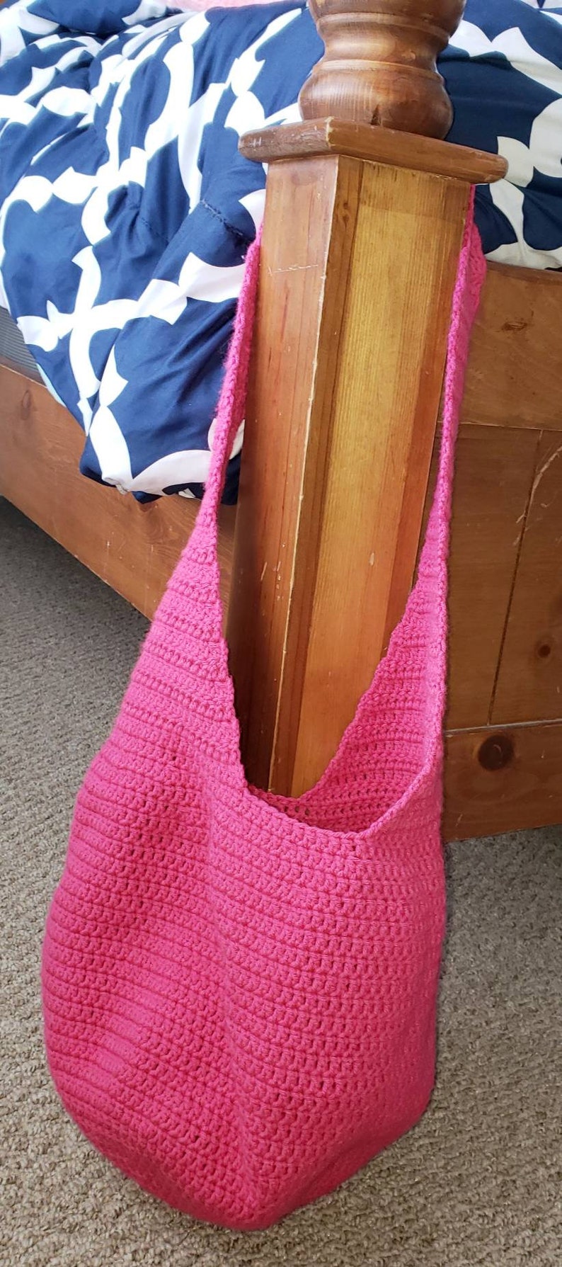 May include: A bright pink, handmade crochet shoulder bag. The bag is a solid color and has a textured appearance. The strap is also crocheted. The bag is shown resting on a carpeted floor next to a wooden bed frame.