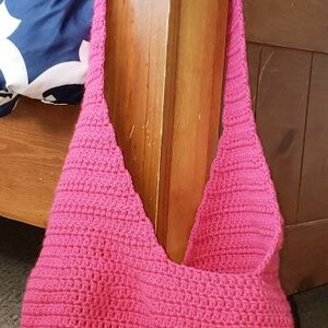 May include: A handmade, hot pink crochet shoulder bag. The bag has a rounded bottom and a wide strap. The bag is hanging on a wooden bed post. The background includes a bed with a blue and white patterned comforter.