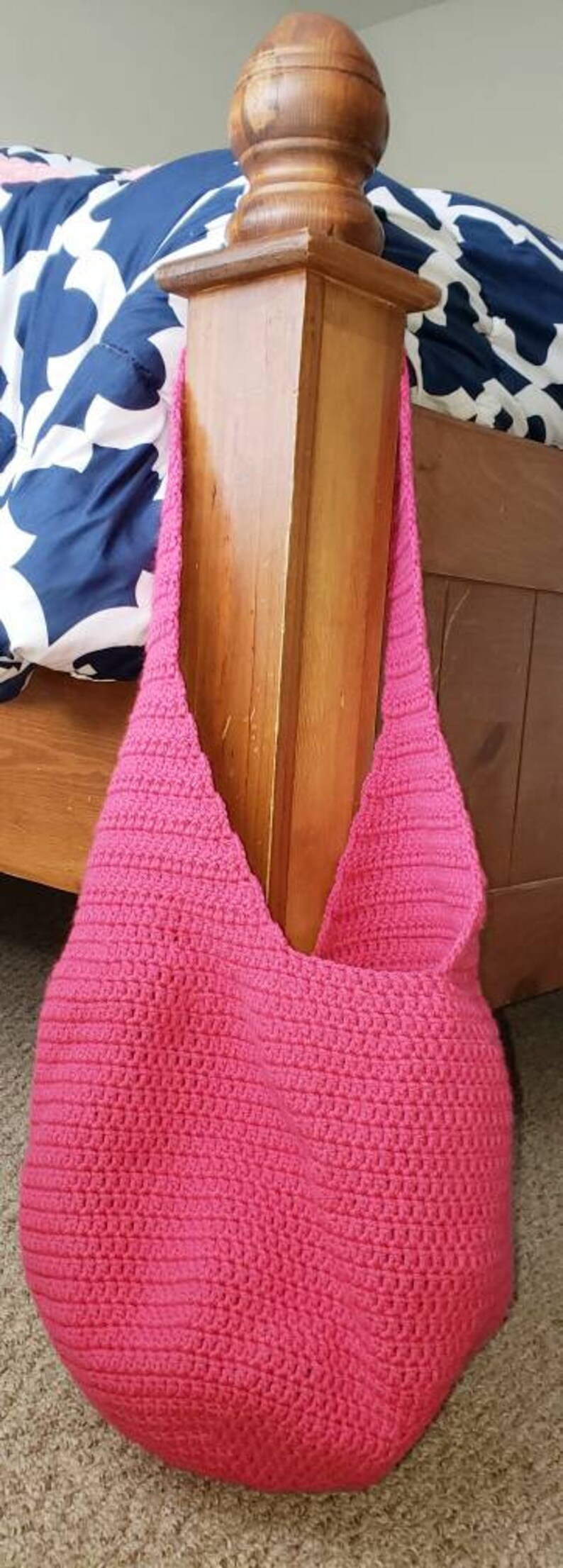 May include: A handmade, hot pink crochet shoulder bag. The bag has a rounded bottom and a wide strap. The bag is hanging on a wooden bed post. The background includes a bed with a blue and white patterned comforter.