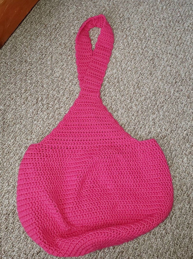 May include: A handmade, bright pink crochet tote bag with a unique teardrop shape. The bag features a long, looped handle and a textured stitch pattern. This stylish bag is perfect for carrying essentials.