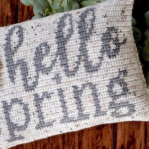 K&ouml;nnte beinhalten: Ein geh&auml;keltes Kissen mit den Worten "Hello Fall" in grauer Schrift auf wei&szlig;em Grund mit Farbflecken. Das Kissen ist mit drei geh&auml;kelten Blumen in Wei&szlig;, Gr&uuml;n und Orange verziert.